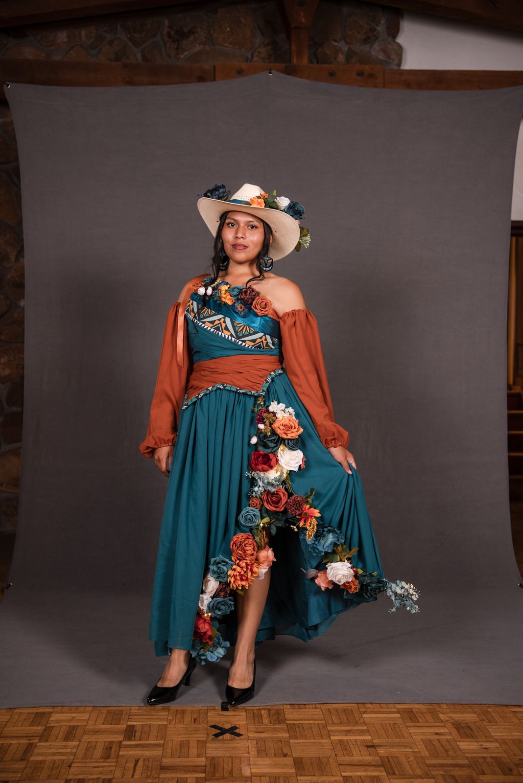 Model in a teal gown with a rust bodice, flowers and a sun hat